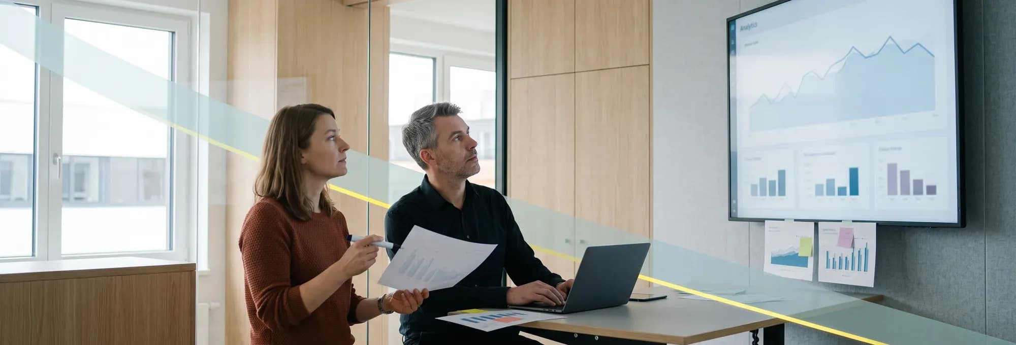 Besprechungsecke in der Verwaltung eines Familienunternehmens. Zwei Kollegen sehen sich einen Ausdruck an, an der Wand hängt ein Bildschirm mit einer Auswertung.